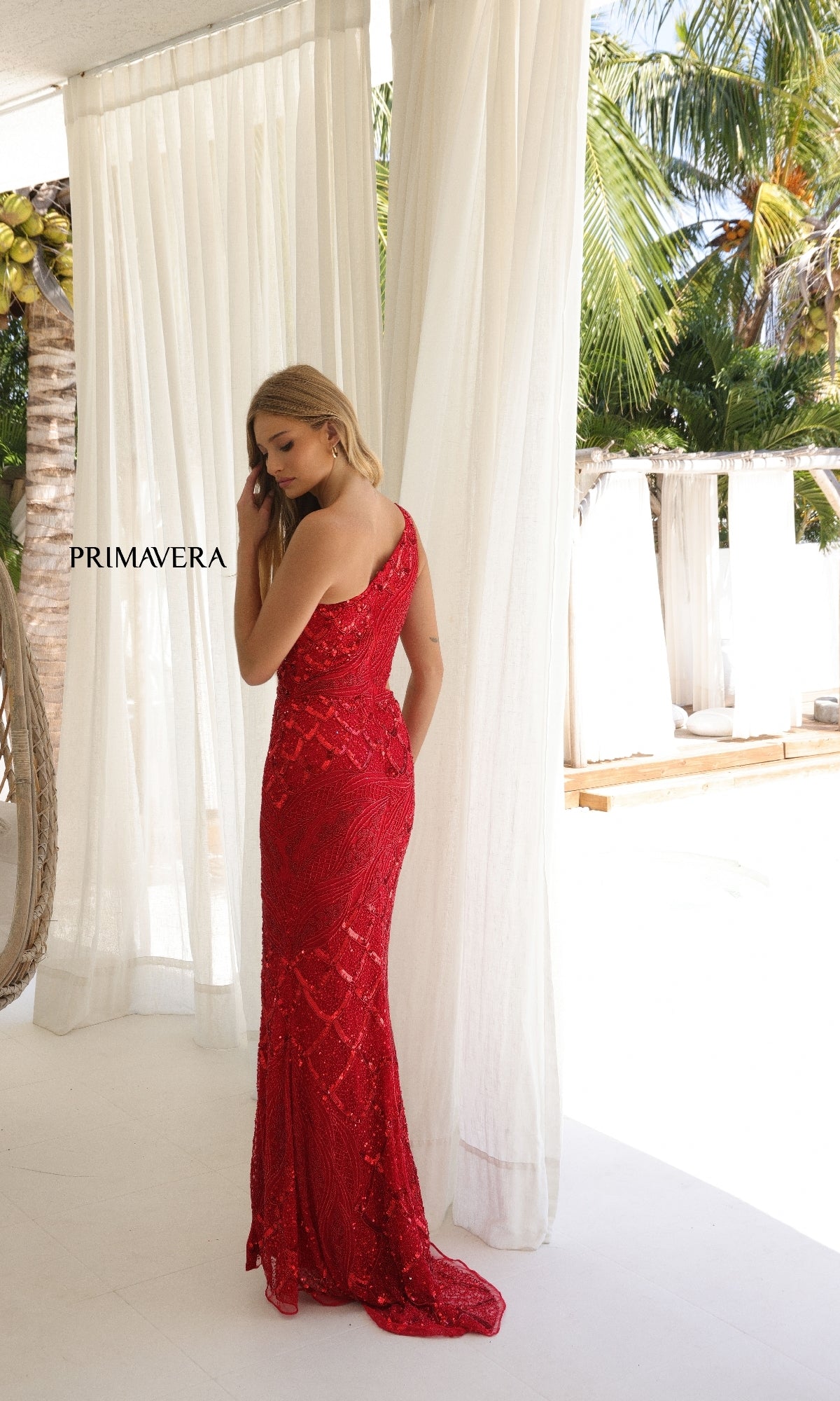 A woman in a red, floor-length sequin gown—Long Prom Dress: Primavera 4329—stands indoors by sheer curtains with palm trees outside.